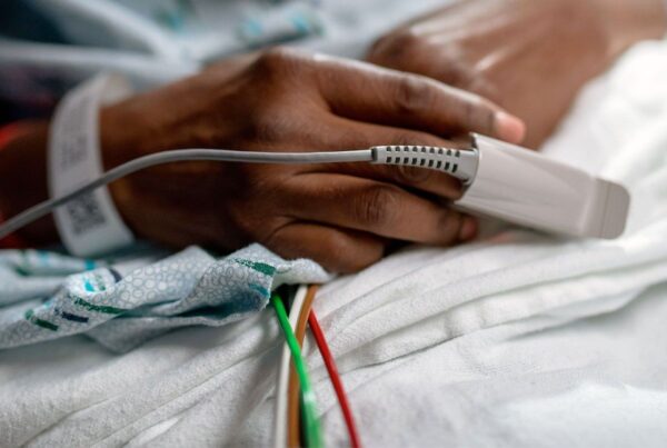 A close-up of a persons hand wearing a hospital wristband, resting on a blanket, with a pulse oximeter clipped to their finger and medical wires visible underneath.
