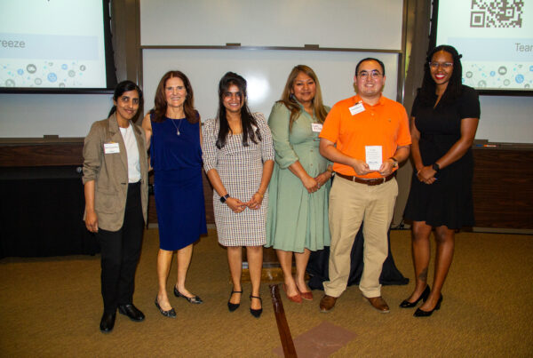 Six people stand indoors in a row, smiling at the camera. They are dressed in business or semi-formal attire, and some wear name tags. A presentation screen and a QR code are visible in the background.